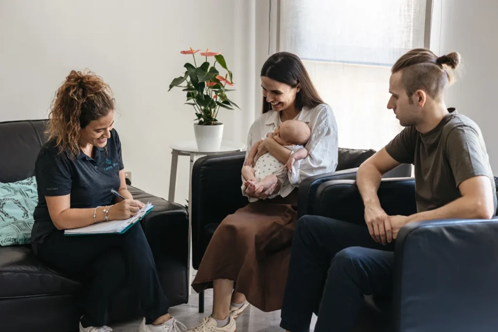 Silvia takes notes while talking with parents and their baby on a sofa.
