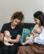 Silvia demonstrates baby positioning with a doll beside a mother holding her baby.
